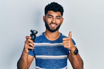 Sticker - Arab man with beard holding electric razor machine smiling happy and positive, thumb up doing excellent and approval sign