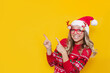 © Марина Демешко - A young smiling blonde woman in a red Christmas sweater, glasses with deer antlers and a red Santa's hat points at copy space for text or design with her fingers isolated on a color yellow background