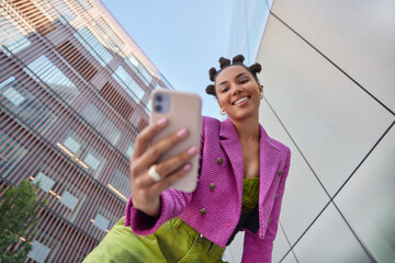 Happy young woman with hairstyle dressed in fashionable clothes makes video call or selfie via smartphone uses mobile app strolls at urban setting films vlog for social networks. Communication