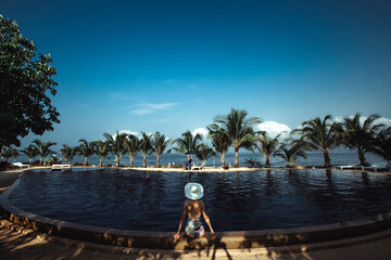 Wall Mural - View from the back of young woman is sitting by the pool on a tropical beach with palm trees.