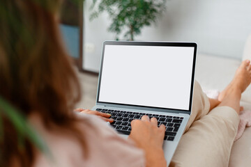 Woman looking at mockup white laptop computer screen on online e learning app web site or moovie at home. Over shoulder close up female hands on keyboard. Woman sit on sofa with laptop screen mock up