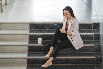 Poster - Cheerful Thai young businesswoman sitting on the stairs working on her tablet in the office