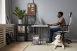 © Seventyfour - Full length portrait of young African-American man working in office with dog laying on floor, pet friendly workspace, copy space
