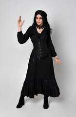 Sticker - Full length portrait of dark haired woman wearing  black victorian witch costume with corset,  standing pose with  gestural hand movements,  against studio background.