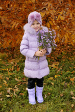 A Beautiful Girl In A Violet Turban On Her Head In A Purple Down Jacket And Boots Stands Against The Background Of An Autumn Yellowed Tree With Lilac Flowers In Her Hands