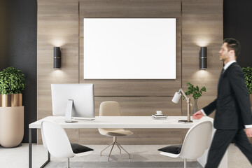 Poster - Businessman walking in modern wooden office interior with empty white mock up poster, desktop, equipment and other items. Workplace and design concept.