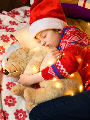  Child girl sleeping in new year or christmas decoration. Holiday lights and gifts, Christmas tree decorated with toys. She's wearing a red sweater and a Santa helper hat