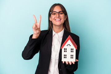 Wall Mural - Young business woman holding a toy home isolated on blue background showing number two with fingers.