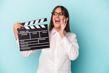 Wall Mural - Young caucasian woman holding clapperboard isolated on blue background shouting and holding palm near opened mouth.