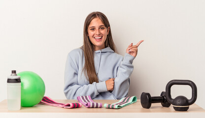 Wall Mural - Young caucasian woman sitting at a table with sport equipment isolated on white background smiling cheerfully pointing with forefinger away.