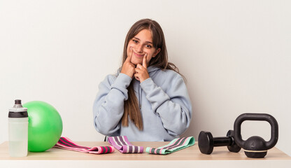 Wall Mural - Young caucasian woman sitting at a table with sport equipment isolated on white background doubting between two options.