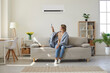 © Studio Romantic - Woman who's sitting on sofa under warm plaid in living room switches off her air conditioner on wall. Young girl adjusting modern AC system, regulating temperature and enjoying cool fresh air at home