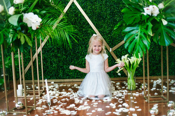  Little cute girl with blond hair in a white dress  and white flowers, lilies and orchids on a  background with a green tropical plants