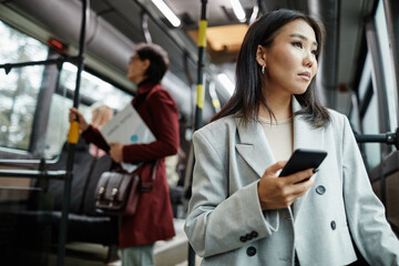 Wall Mural - Portrait of elegant Asian woman looking at window on bus while traveling by public transport and holding smartphone, copy space