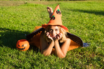 adult woman with blonde hair and a bit chubby in the park enjoying the halloween party. The woman is wearing black lingerie with black wings, an orange witch's hat and a pumpkin to collect candy. 