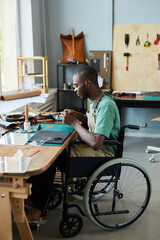 Vertical full length portrait of young African-American man in wheelchair making handmade bag in leatherworkers workshop