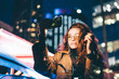 Curly haired young woman in glasses wearing light coat listens to music with headphones and makes selfie using phone camera against skyscraper in night city.