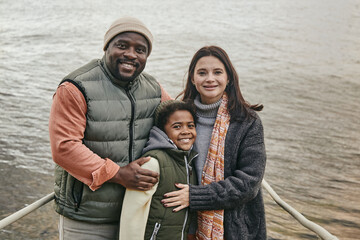 Wall Mural - Portrait of multiethnic family embracing their son and smiling at camera while standing outdoors against the lake