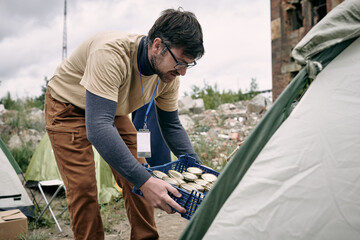 Wall Mural - Male volunteer with box of canned food bending by tent where migrants live