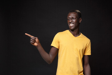 Wall Mural - Smiling young african american man guy in yellow t-shirt posing isolated on black wall background studio portrait. People emotions lifestyle concept. Mock up copy space. Pointing index finger aside.