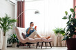 Happy calm Caucasian woman relax sit in comfort chair in cozy home interior use smartphone. Smiling young woman use mobile phone drink cup of coffee or tea, have rest in armchair with home plants.