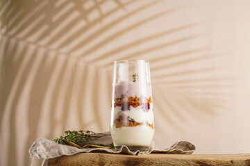 Wall Mural - Tall glass with a layered creamy yoghurt dessert trifle with cookies, amaretti, cherries and thyme on a linen cloth on a rustic wooden table at a tropical setting