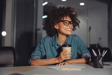 Wall Mural - Smiling young multiethnic lady sitting at work desk and holding cup of coffee in the office