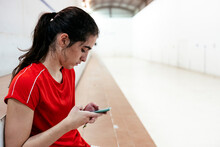 Side View Of A Female Player Of A Traditional Spanish Game "Valencian Pelota" Using Phone Before Play Game Sitting On Bleachers Of Indoor Court