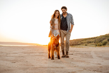 Wall Mural - Young white couple hugging and smiling while walking with their dog