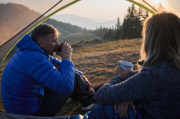Wall Mural - View from inside tent of man and woman sitting inside tent and enjoying pleasant taste of coffee brewed in fresh air. Recreation in tent set up on mountain hill during long hiking.