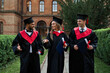 Three smiling gradutes international friends in graduation gowns walking in campus
