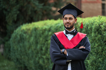 Serious indian graduate in graduation robe with crossed arms looking forward, copy space for text