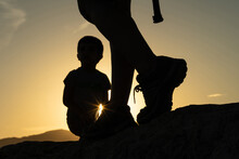 Silhouette Of The Legs Of A Woman Doing Trekking In The Mountain With Her Son And The Sun Creating A Sun Star At Sunset