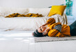 © hitdelight - Woman in casual clothing with headphones lying down the floor in her living room and enjoys listening the music. Leisure time of single woman relaxing in isolation streaming songs from player.