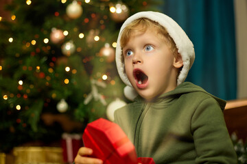 Wall Mural - Cute boy in a Christmas hat opens a gift under a decorated Christmas tree. Happy boy with his mouth open in surprise holds a magic glowing box in his hands.