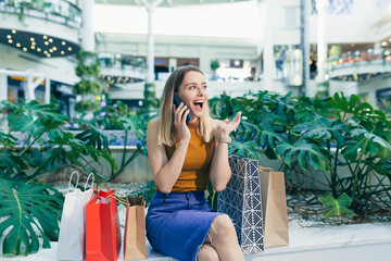 Wall Mural - Young woman consumer in the mall browses chat and uses using a smartphone. female standing with a mobile phone in her hands in shopping center. indoor. happy shopper girl with gift bags make purchases