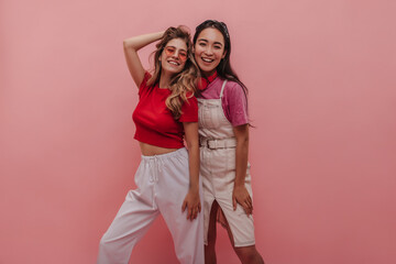 Wall Mural - Caucasian and ukrainian young school girl friends posing for parents on summer vacation. Blonde with wavy hair and red crop top with girl in denim dress and pink shirt. Lifestyle female beauty concept