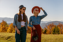 Outdoor Fashion Portrait Of Two Young Confident Women Wearing Stylish Autumn Outfits With Hats, Sunglasses, Bags, Denim Clothes, Posing, Walking In Mountains. Copy, Empty Space For Text