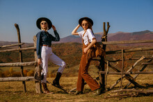 Two Fashionable Models Wearing Stylish Autumn Outfits With Trendy Hats, Sunglasses, Bags, Cowboy Boots, Posing In Mountains. Full Length Outdoor Portrait. Copy, Empty Space For Text