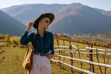 Outdoor Fashion Portrait Of Confident Blonde Woman Wearing Stylish Sunglasses, Hat, Blue Denim Shirt, White Jeans, With Brown Suede Shopper Bag,  Posing In Autumn Mountains. Copy, Empty Space For Text