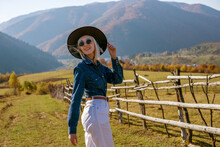 Happy Smiling Fashionable Woman Wearing Stylish Sunglasses, Black Wide Brim Hat, Blue Denim Shirt, White High Waist Jeans, Posing, Walking In Autumn Mountains. Copy, Empty Space For Text