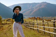 Fashionable Woman Wearing Stylish Sunglasses, Black Wide Brim Hat, Blue Denim Shirt, White High Waist Jeans, Posing In Autumn Mountains. Copy, Empty Space For Text