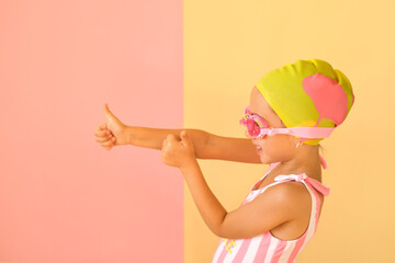 Cheerful positive girl in a trendy striped swimsuit, Swimming Goggles and a pool cap  showing success gesture, thumb up with hand , saying well done. Pink-yellow studio background. Super cool swimming