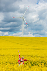 Sticker - Woman in red and white striped jacket with wind turbine on background in rapeseed field