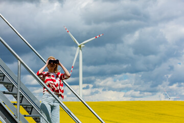 Sticker - Woman in red and white striped jacket watching in binoculars with wind turbine on background in rapeseed field