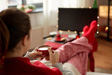 Poster - christmas, winter holidays and leisure concept - close up of young woman watching tv and drinking coffee with her feet on table at cozy home