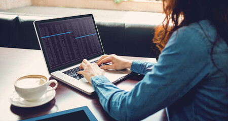 Caucasian female programmer sitting at desk with coffee, using laptop with coding on screen