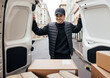 © Artem Varnitsin - Smiling man in uniform and cap looking at cardboard boxes while standing at van trunk