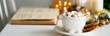 Long Banner Christmas Light Gray Background With Copy Space. Reading Book On Cozy Winter Evening With Candles, Cocoa Hot Chocolate With Marshmallows And Cookies. Side View Of Table, Chair In Home