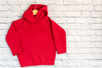 Wall Mural - Red kids sweatshirt with hood with clothes hanger on white well brick background. Fashionable unisex clothing, hoodie, casual youth style, sports. Blank hoody mock up